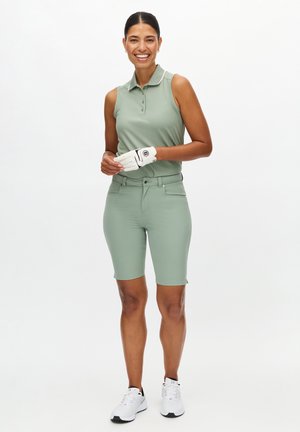 Polo sin mangas de color verde claro con cuello, combinado con pantalones cortos ajustados a juego. Guante de golf blanco en la mano izquierda, zapatillas blancas.