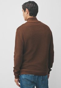 Brauner gestrickter Pullover mit hohem Kragen, strukturiertem Material und langen Ärmeln. Kombiniert mit blauen Jeans, die Details der Gesäßtaschen zeigen.