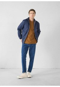 Giacca a vento blu navy con zip frontale, t-shirt marrone, jeans azzurri e sneakers beige. Tessuto liscio con un design semplice.