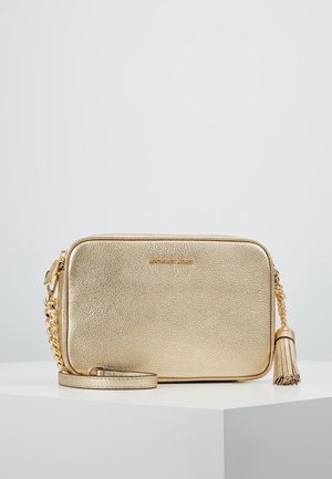 Gouden leren Michael Kors crossbodytas met kettingband en kwastdetail, geplaatst op een wit oppervlak tegen een lichtgrijze achtergrond.