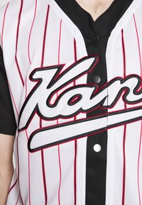 Maglietta da baseball bianca con strisce verticali rosse e maniche nere. Presenta un grande testo ricamato in nero e due bottoni sul davanti.