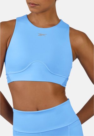 Femme athlétique portant un crop top bleu clair à col montant et des leggings taille haute assortis, debout, les mains sur les hanches.