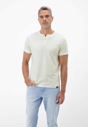 Mann steht mit der Hand in der Tasche, trägt ein cremefarbenes Henley-Shirt mit kurzen Ärmeln und hellblaue Jeans vor einem weißen Hintergrund.