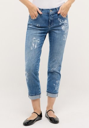 Frau trägt blaue, umgeschlagene Jeans mit weißer Blumen- und Schleifenstickerei, Hände in den Taschen und schwarze lackierte Mary-Jane-Schuhe.