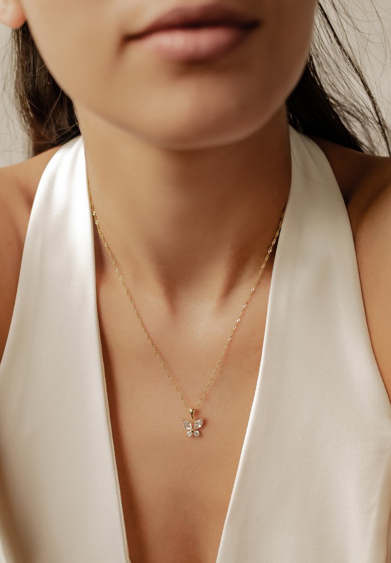 Collana d'oro con ciondolo a forma di farfalla decorata con gemme trasparenti. La farfalla ha due ali e un corpo centrale, poggiato sopra una maglietta bianca.