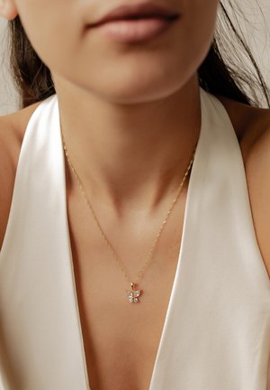 Collier en chaîne dorée avec un petit pendentif en forme de papillon porté au cou d'une femme vêtue d'un haut blanc sans manches.