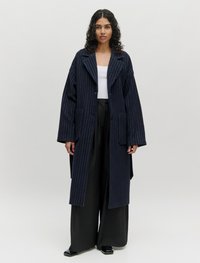 Navy gestreepte jas met oversized silhouet, brede mouwen en twee voorzakken, gelaagd over een witte top en zwarte wijde broek.