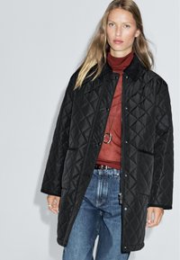 Gepolsterte schwarze Jacke mit Druckknopfverschluss, ausgestattet mit einem Kragen und zwei Vorderaschen, getragen über einem roten Rollkragenpullover und Jeans.