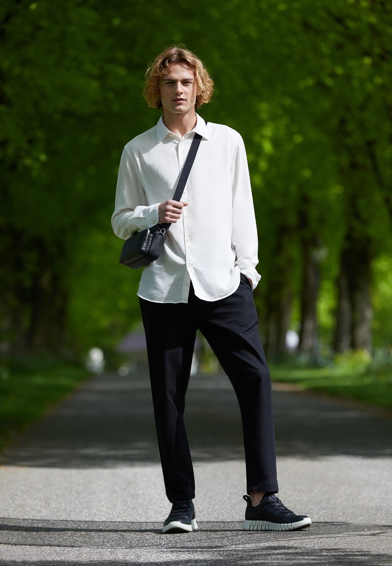 Vit långärmad skjorta, svarta byxor och svarta sneakers. Svart axelremsväska. Modellen står på en stig omgiven av grön bebyggelse.
