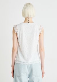 Hvit blondebluse med blomstermønster og bølgete skulderdetaljer, kombinert med lyseblå denimbukser. Bakview viser intrikat blonde tekstur.