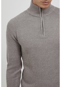 Pull en tricot gris avec col montant et demi-fermeture éclair. Présente un motif texturé et des manches longues, fabriqué dans un matériau doux et léger.
