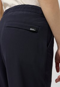 Pantalon extensible navy avec taille élastique, doté d'une poche au dos et d'une étiquette de marque. Tissu lisse avec une finition mate.