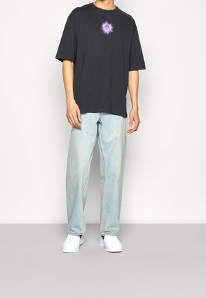 Black oversized t-shirt with a purple graphic design, paired with light blue straight-leg jeans and white sneakers. Light background.