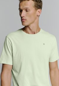 Lichtgroene katoenen T-shirt met een ronde hals, korte mouwen en een klein verticaal streepjespatroon op de linkerborst.