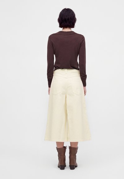 Personne aux cheveux courts et foncés portant un haut marron à manches longues, un pantalon crème à larges jambes coupé au-dessus de la cheville, et des bottines marron, vue de dos.