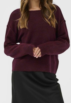 Frau trägt einen locker sitzenden bordeauxfarbenen Langarmpullover mit gerippten Bündchen und einen schwarzen Rock, die Hände an der Taille verschränkt, vor einem schlichten Hintergrund.