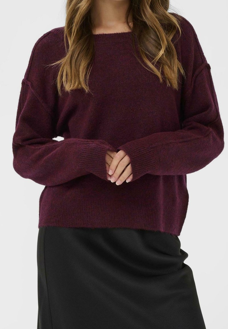 Femme portant un pull bordeaux ample à manches longues avec des poignets côtelés et une jupe noire, les mains jointes à la taille, sur un fond uni.