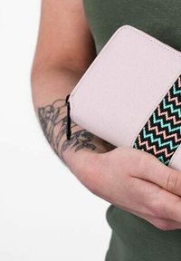 Růžová peněženka z látky s tkaným vícebarevným zigzag vzorem. Obsahuje stahovací zip a texturovaný povrch, držena proti zelenému oděvu.