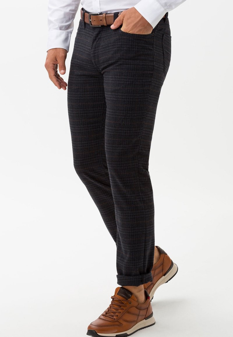 Homme portant un pantalon à carreaux foncés avec revers roulés, ceinture en cuir marron, chemise blanche et baskets marron, debout avec une main dans la poche.