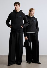 Μαύρες φούτερ με κουκούλα και εμπρόσθιες τσέπες, διακοσμημένες με κεντημένο λογότυπο. Τα μοντέλα φορούν φαρδιά μαύρα παντελόνια και κρατούν μια μικρή μαύρη τσάντα.