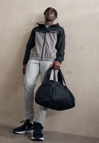 Veste de sport noire et grise, avec fermeture éclair à l'avant, taille élastique, pantalon de jogging gris, sac de sport noir et baskets noires avec accents blancs.
