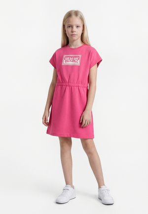 Dziewczyna stoi w różowej sukience Versace z elastycznym paskiem w talii i białych trampkach, na jednolitym białym tle.