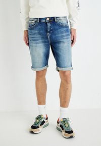Shorts en denim bleu clair, avec des ourlets roulés, design à cinq poches, fermeture par bouton en cuivre. Associés à des baskets colorées et des chaussettes blanches mi-mollet.