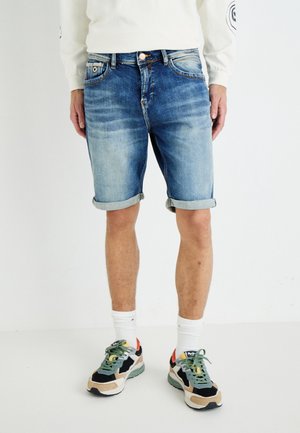 Shorts en denim bleu clair, avec des ourlets roulés, design à cinq poches, fermeture par bouton en cuivre. Associés à des baskets colorées et des chaussettes blanches mi-mollet.