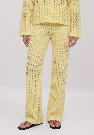 Gelbe gehäkelte Strickhose und passender geknöpfter Cardigan mit lockerem Sitz, getragen mit schwarzen Flip-Flops mit roten Riemen.