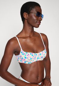 Top bikini floreale con fiori rosa, blu e gialli su uno sfondo bianco. Presenta sottili spalline bianche e un tessuto liscio e leggero.