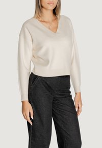 Maglione corto color crema con scollatura a V realizzato in un tessuto morbido, abbinato a pantaloni neri a vita alta. Le maniche sono leggermente a palloncino, aggiungendo volume.