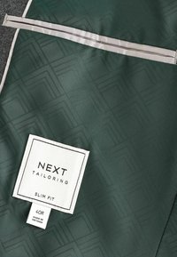 Material textil verde cu un model geometric texturat, având o etichetă albă care scrie "NEXT TAILORING SLIM FIT" și detalii despre mărime.