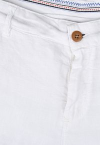 Bílé lněné šortky mají knoflíkové zapínání s hnědým knoflíkem. Pás má pruhovaný vzor v modré a červené barvě.