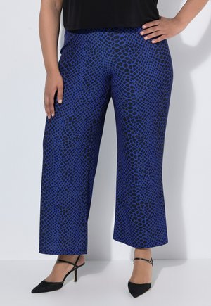 Vrouw draagt blauw-zwarte brede broek met patroon en zwarte sandalen met hakken en bandjes, staat met één hand op de heup en één hand omlaag.