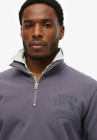Grijze zip-up sweatshirt met een groen gestreepte kraag en "SUPERDRY Athletics" in donkergroen gedrukt op de borst. Soepele textuur.