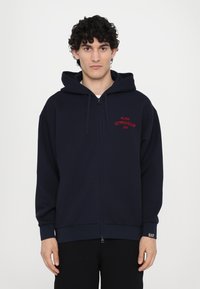 Marinblå zip-up hoodie med en framficka, som har röd brodering "MILANO GYMNASIUM EA7", samt en huva med dragsnodd. Mjuk tygkvalitet, avslappnad passform.