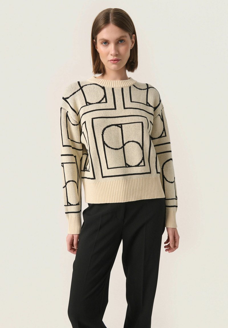 Pulover tricotat de culoare crem cu modele geometrice îndrăznețe în negru. Guler rotund, manșete și talie ribbate. Asortat cu pantaloni negri.