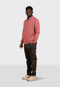Polo rouge à manches longues avec fermeture éclair quart et écusson, associé à un pantalon sombre et des baskets beiges. Texture lisse et coupe décontractée.