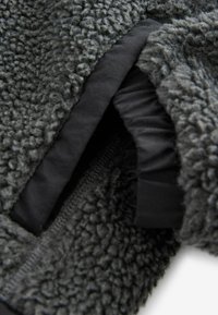 Tessuto in pile grigio, testurizzato, con una superficie morbida e pelosa; presenta un rivestimento elasticizzato nero e dettagli con chiusura a zip.