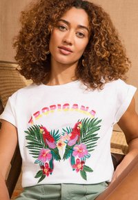 T-shirt en coton blanc avec broderie multicolore de perroquets et de fleurs tropicales, avec le mot "TROPICANA" en haut en couleurs dégradées.