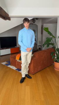 Homme portant une chemise bleu clair, un pantalon beige et des chaussures noires, debout dans un salon avec un canapé couleur rouille et une grande plante verte.