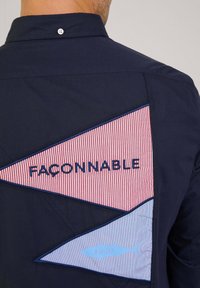 Marineblauw shirt met driehoekige patches in rode en blauwe strepen, met de tekst "FAÇONNABLE" en een visgrafiek met "1950" in lichtblauw.