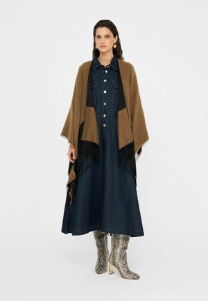 Poncho marron et noir avec des franges sur une robe en denim, arborant des boutons et des manches longues, associé à des bottes montantes en simili cuir serpent brillantes.