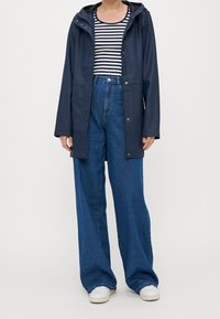 Tmavě modrý nepromokavý plášť s kapucí, doplněný pruhovaným topem a volnými modrými džínami. Celý outfit doplňují bílé tenisky.