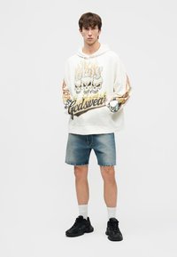 Noor mees, kes kannab valget kapuutsiga pusa, millel on kolju ja leegi motiiv, teksapükse, valgeid sokke ja musti tossusid, seisab käed taskutes.