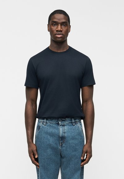 Man wearing a plain black short-sleeve t-shirt tucked into high-waisted blue jeans, standing against a white background.