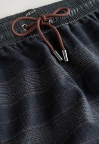 Donkerblauwe, textuurshorts met een subtiel geruit patroon. Beschikt over een elastische tailleband met een bruine trekkoord en metalen uiteinden. Merkenlabel zichtbaar.