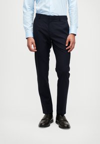 Mornarske hlače ravnog kroja, izrađene od glatke tkanine. Imaju ravni prednji dio, petlje za remen i blagi sjaj. Uparene su s crnim poslovnim cipelama.