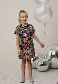 Schwarzes Blumenkleid mit bunten Pailletten und kurzen Ärmeln, kombiniert mit funkelnden pinken Schuhen. Der Hintergrund zeigt silberne Discokugeln.