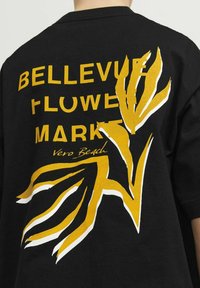 Juodas marškinėliai su ryškiu geltonu tekstu "BELLEVUE FLOWER MARKET" ir stilizuota augalų grafika; minkšta medžiaga, laisvas kirpimas, trumpomis rankovėmis.